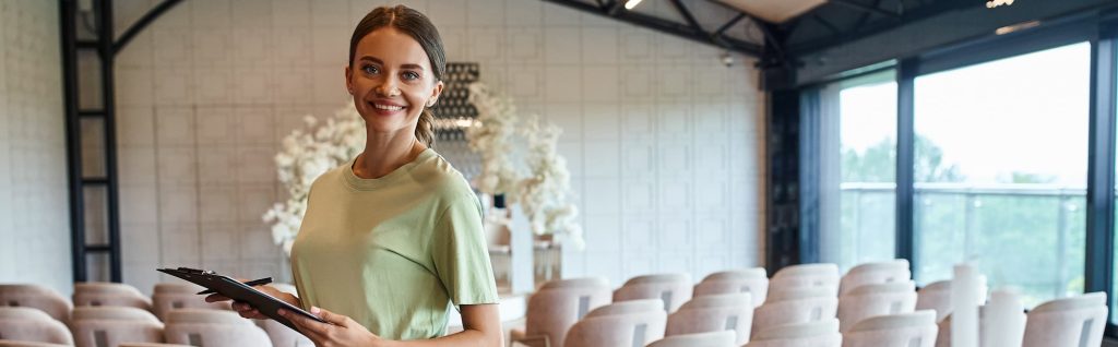 joyful event coordinator with clipboard smiling at camera in decorated banquet event hall, banner