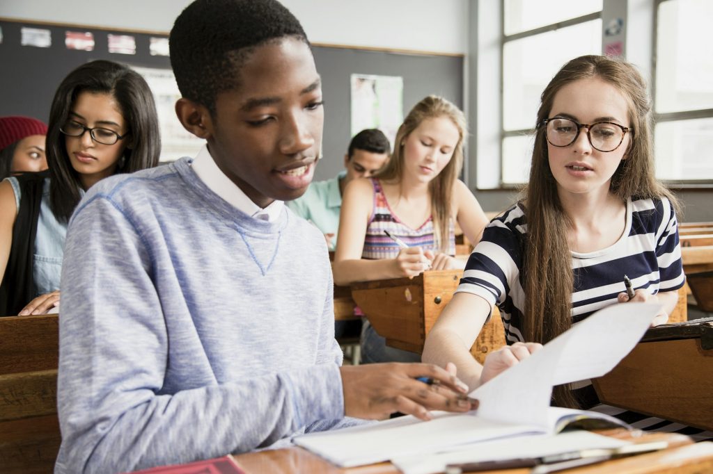 Two students sitting at desk in classroom, helping each other with work